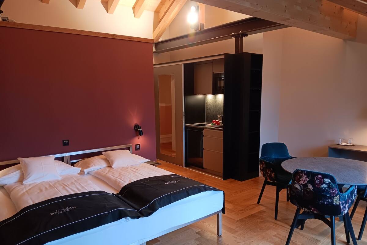 A bedroom with a bed, a desk, and a chair. There is a kitchenette in the background.