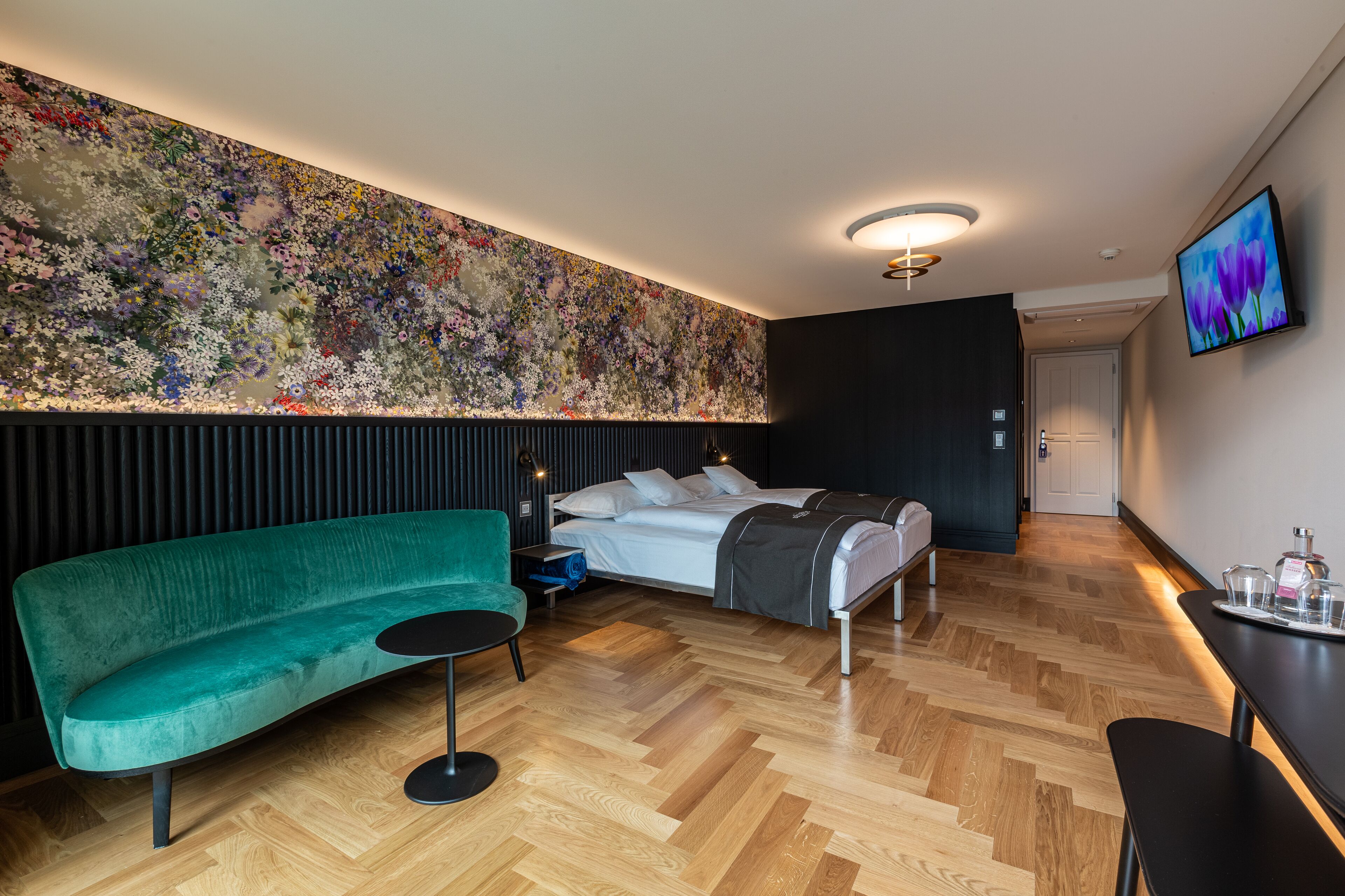Una habitación de hotel moderna con un gran mural floral, un sofá verde, una cama con manta gris y una mesita con bandeja.