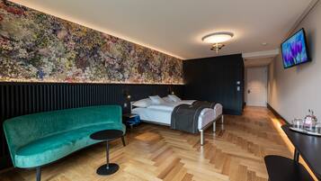 Una habitación de hotel moderna con un gran mural floral, un sofá verde, una cama con manta gris y una mesita con bandeja.