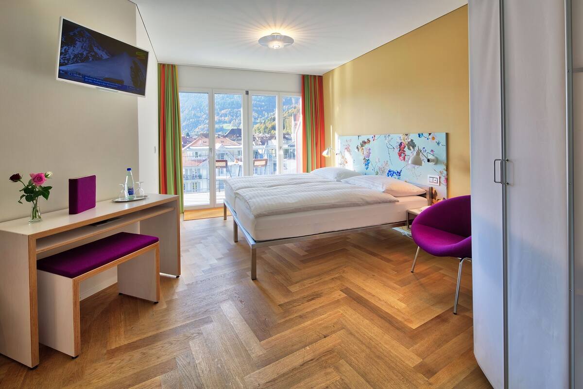 A modern hotel room with a bed, desk, chair, and a large window offering a view of the mountains.