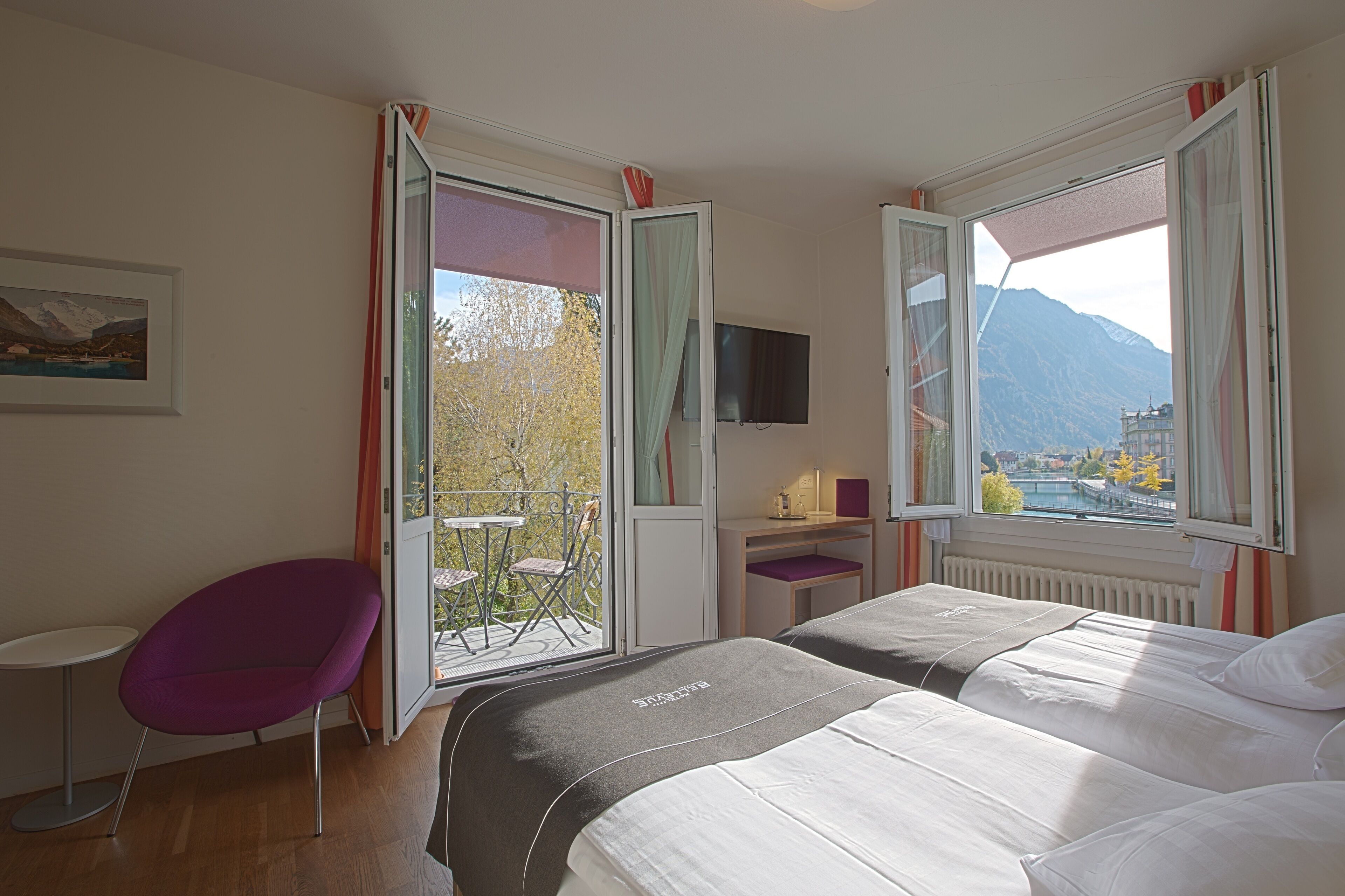 A hotel room with two beds, a desk, a chair, and a balcony with a view of mountains and a lake.