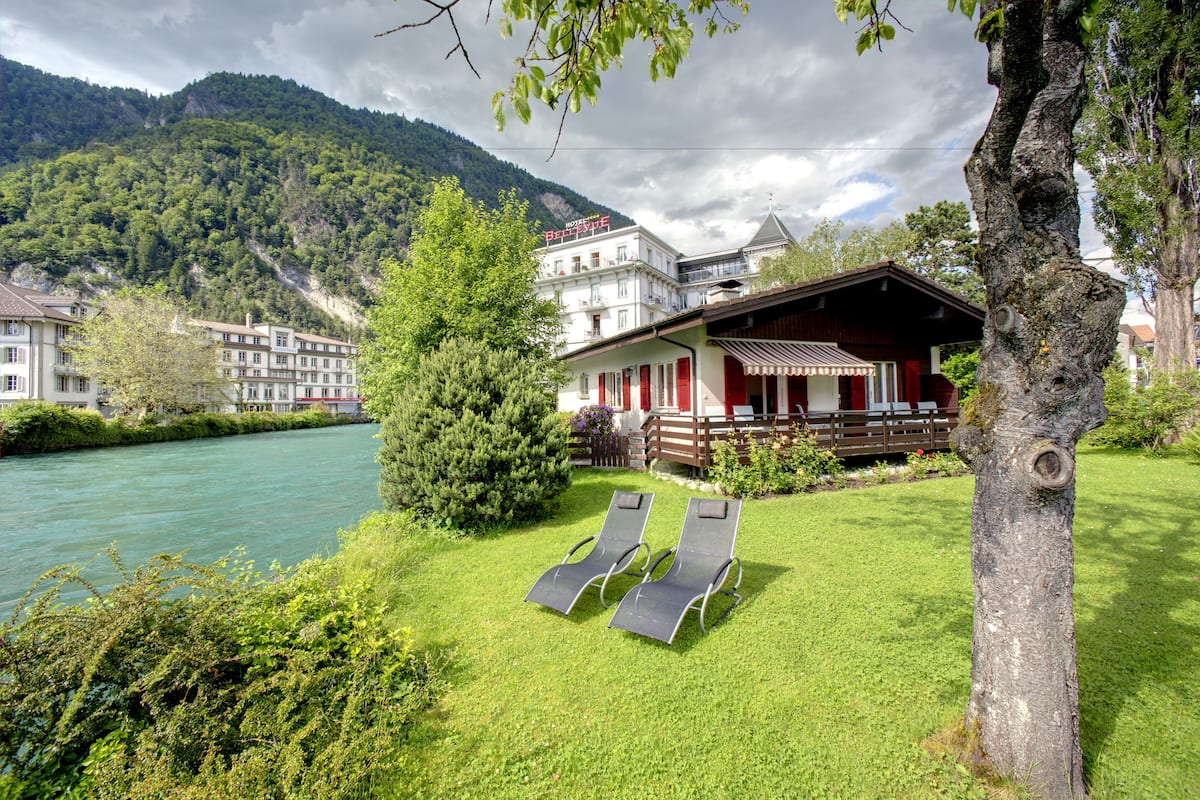 A hotel with a river view, a pool with lounge chairs, and a mountain backdrop.