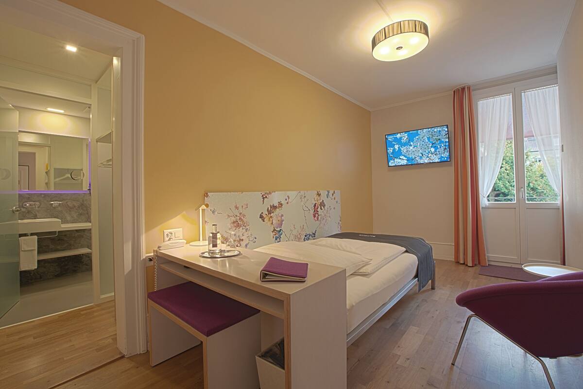 A bedroom with a bed, desk, chair, TV, and bathroom visible through an open door.