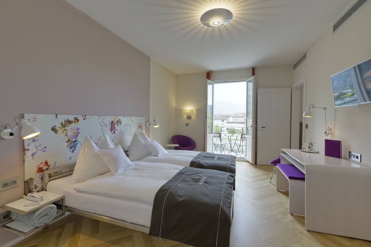 A hotel room with two beds, a desk, a chair, and a balcony with a view.