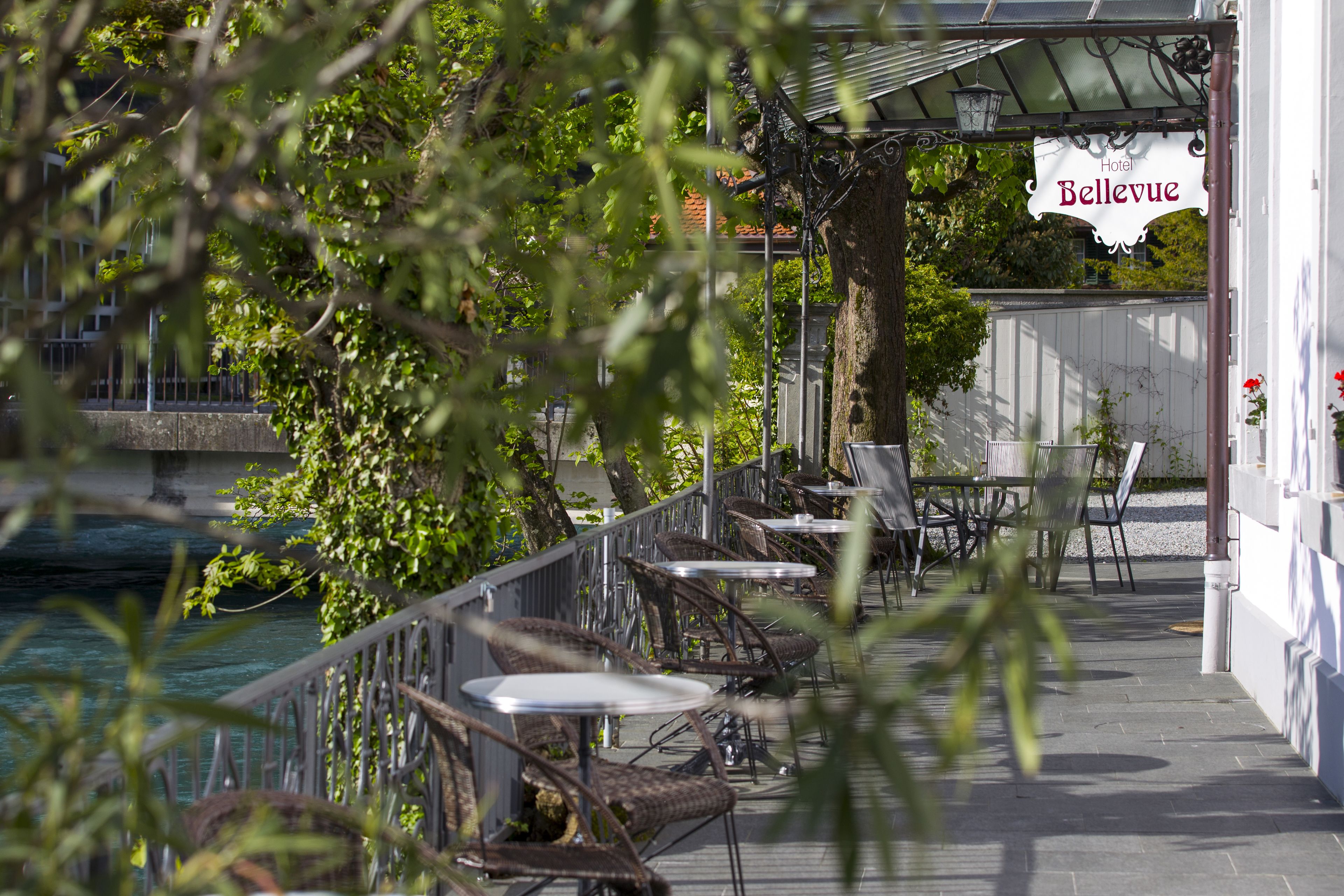 Un espace café en plein air avec des chaises et des tables en osier, une enseigne indiquant « Hôtel Bellevue », et une vue sur une étendue d’eau.