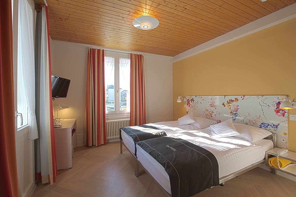 A hotel room with two beds, a TV, a desk, and a window with curtains.