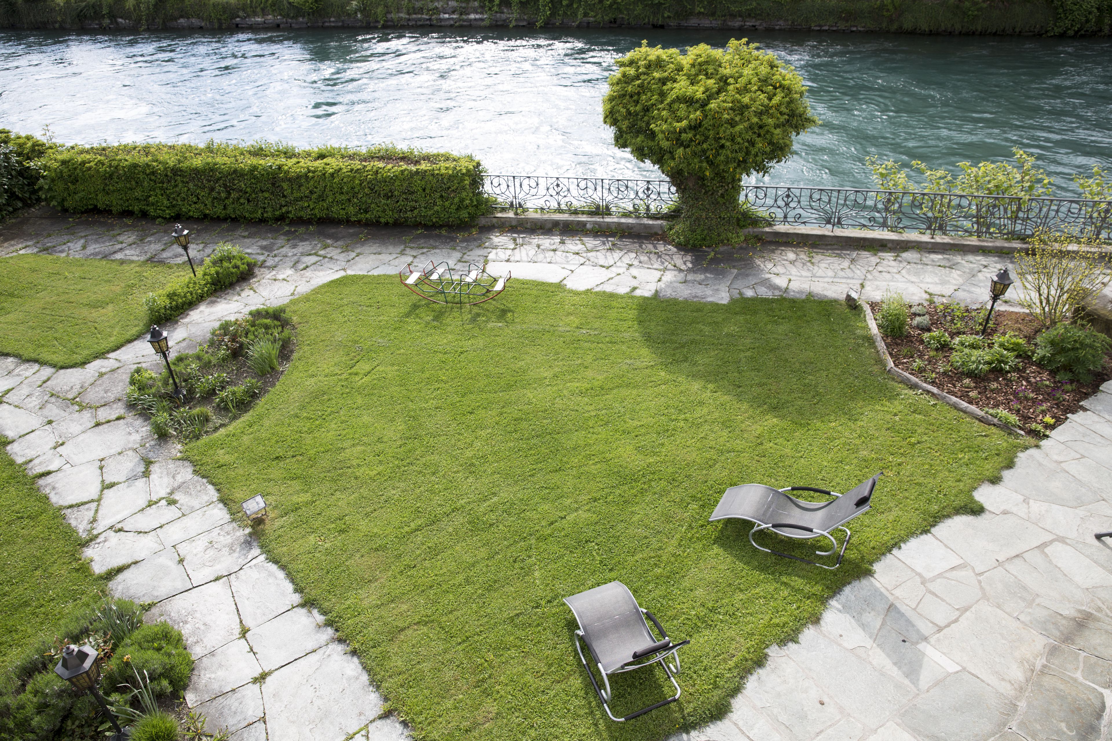 Un patio con césped, dos sillones de descanso, un árbol pequeño y un río al fondo.