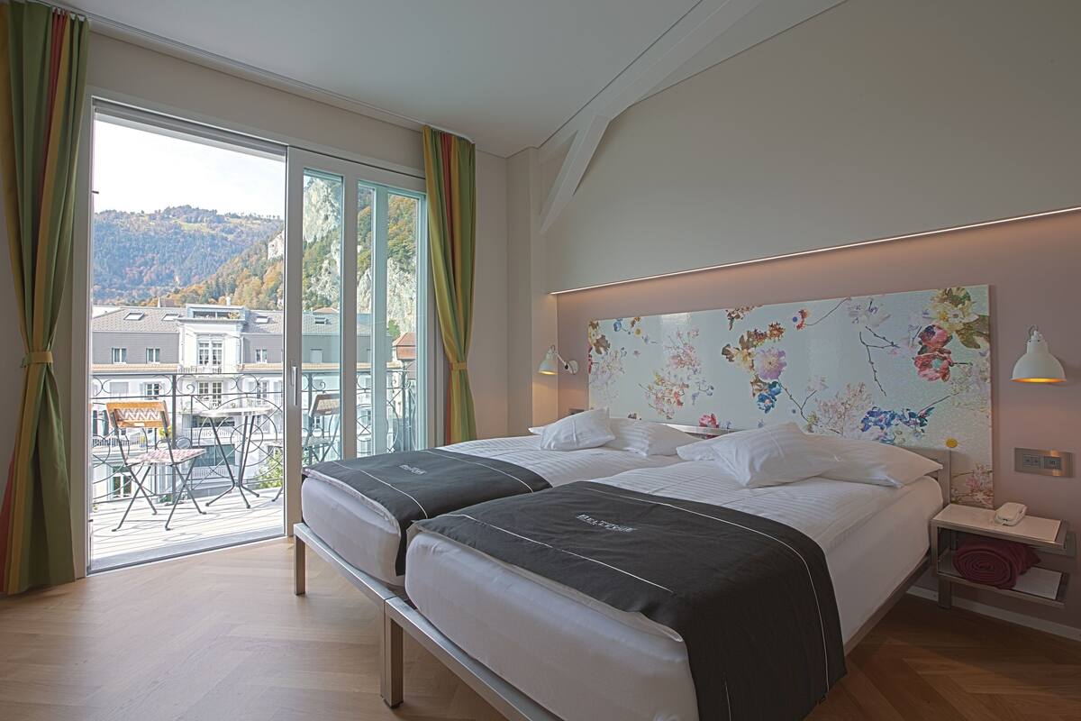 A hotel room with a large bed, a balcony with a view of buildings and hills, and a floral headboard.