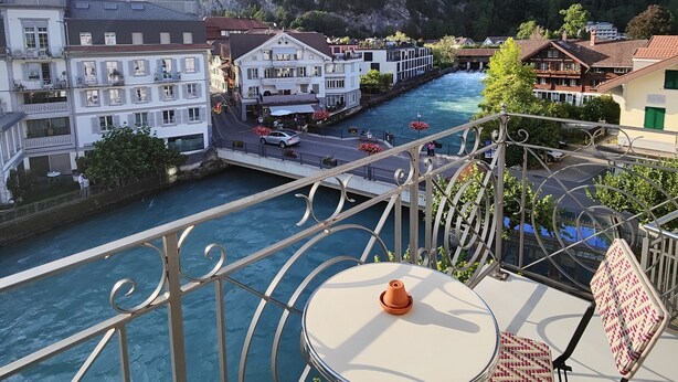 A balcony with a table and chairs overlooking a river, with buildings and a mountain in the background. - Boutique Hotel Bellevue (Interlaken)