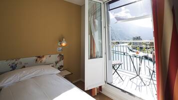 A hotel room with a bed, a nightstand, a lamp, and a balcony with a view of mountains and a body of water.