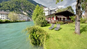 A riverside hotel with a view of a mountain, a wooden deck with lounge chairs, and a building with red shutters. - Boutique Hotel Bellevue (Interlaken)