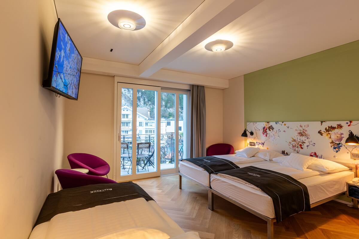A hotel room with two beds, a TV, a balcony with a view of buildings, and a floral wall design.