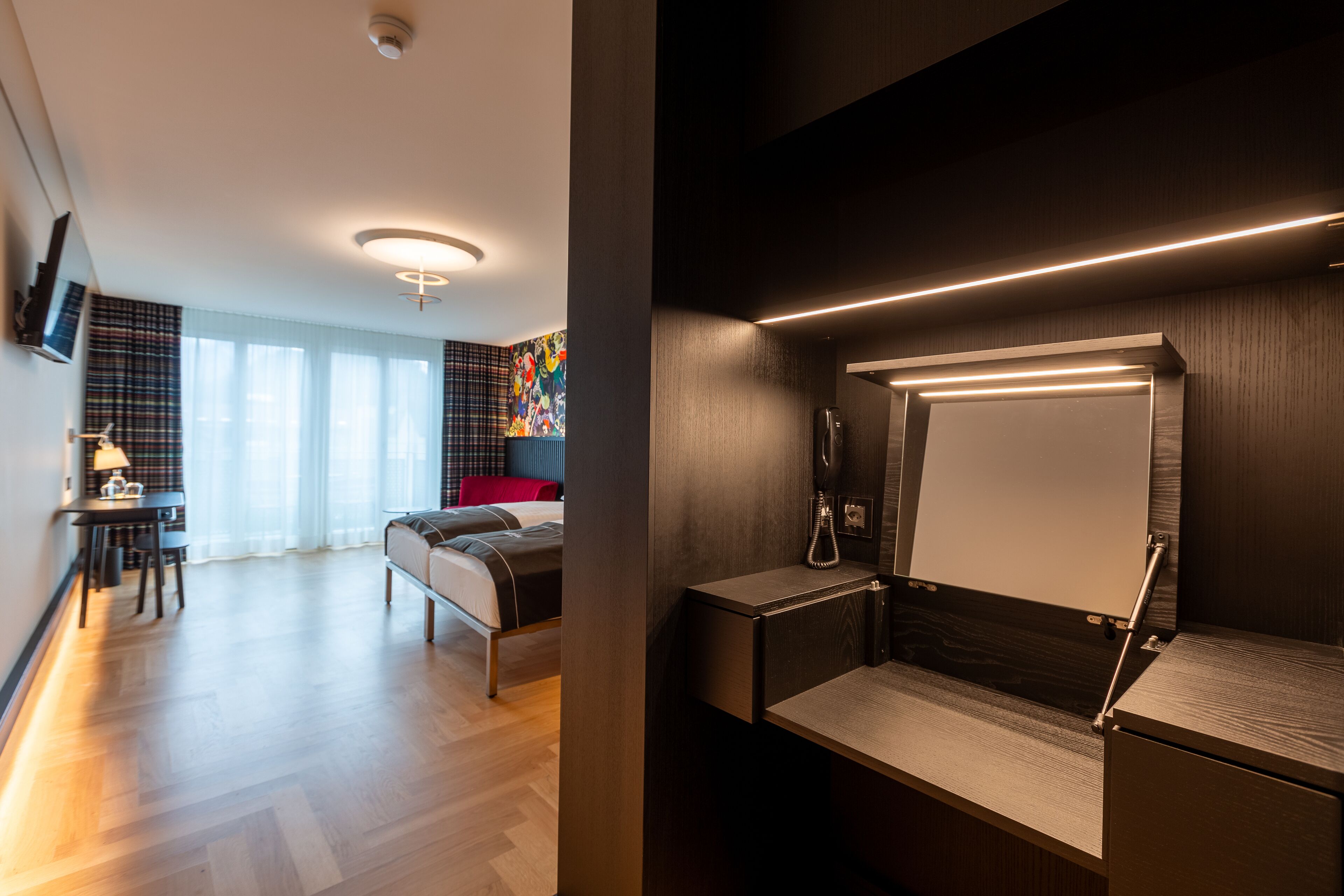 Ein modernes Hotelzimmer mit Holzboden, einem Bett, einem Schreibtisch mit Lampe und einem großen Fenster mit Vorhängen.