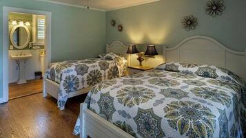 A bedroom with two beds, a ceiling fan, a nightstand with lamps, and a bathroom with a mirror and sink.