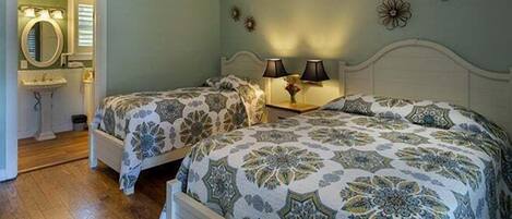 A bedroom with two beds, a ceiling fan, a nightstand with lamps, and a bathroom with a mirror and sink.
