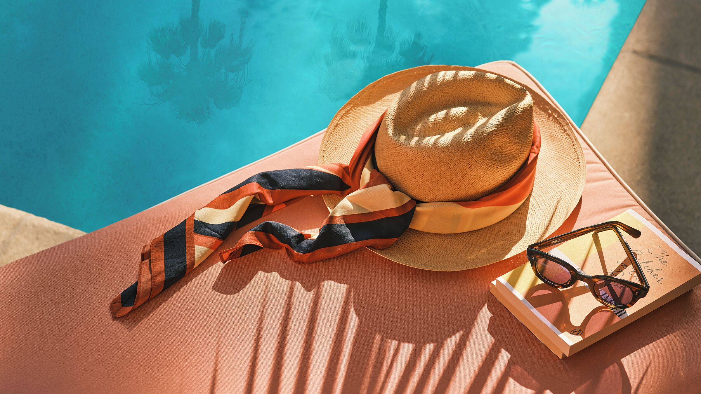 Una tumbona con sombrero, gafas de sol y un libro junto a la piscina.