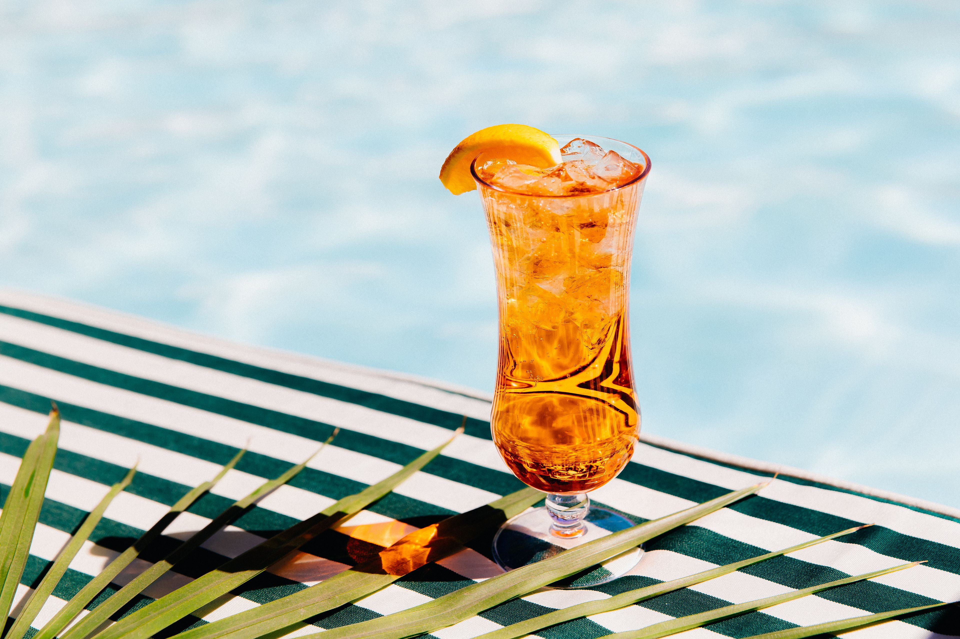 A tall glass with a lemon slice garnish, placed on a striped beach chair by a pool.