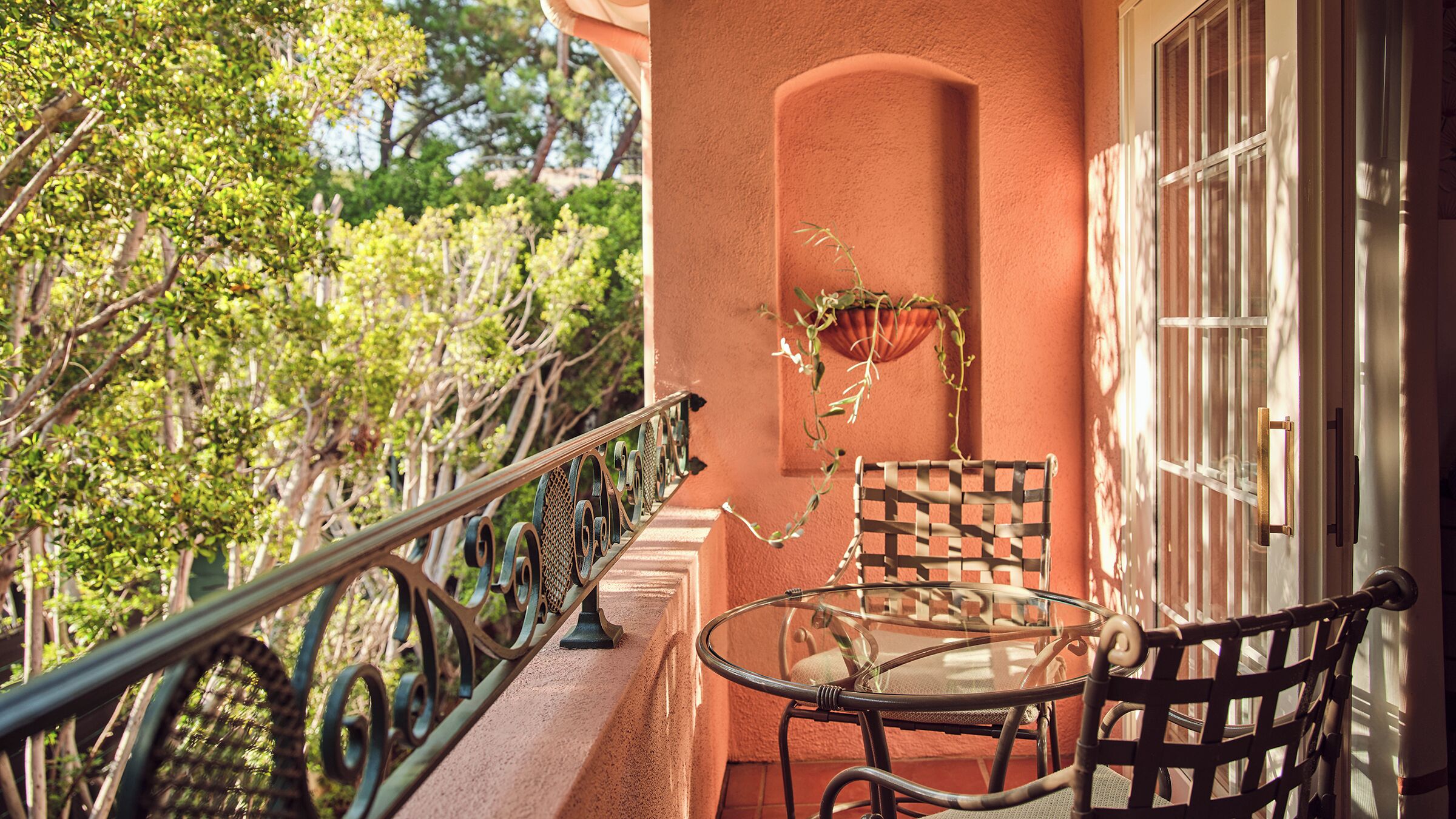 A balcony with a glass table and chairs, a potted plant, and a view of trees.