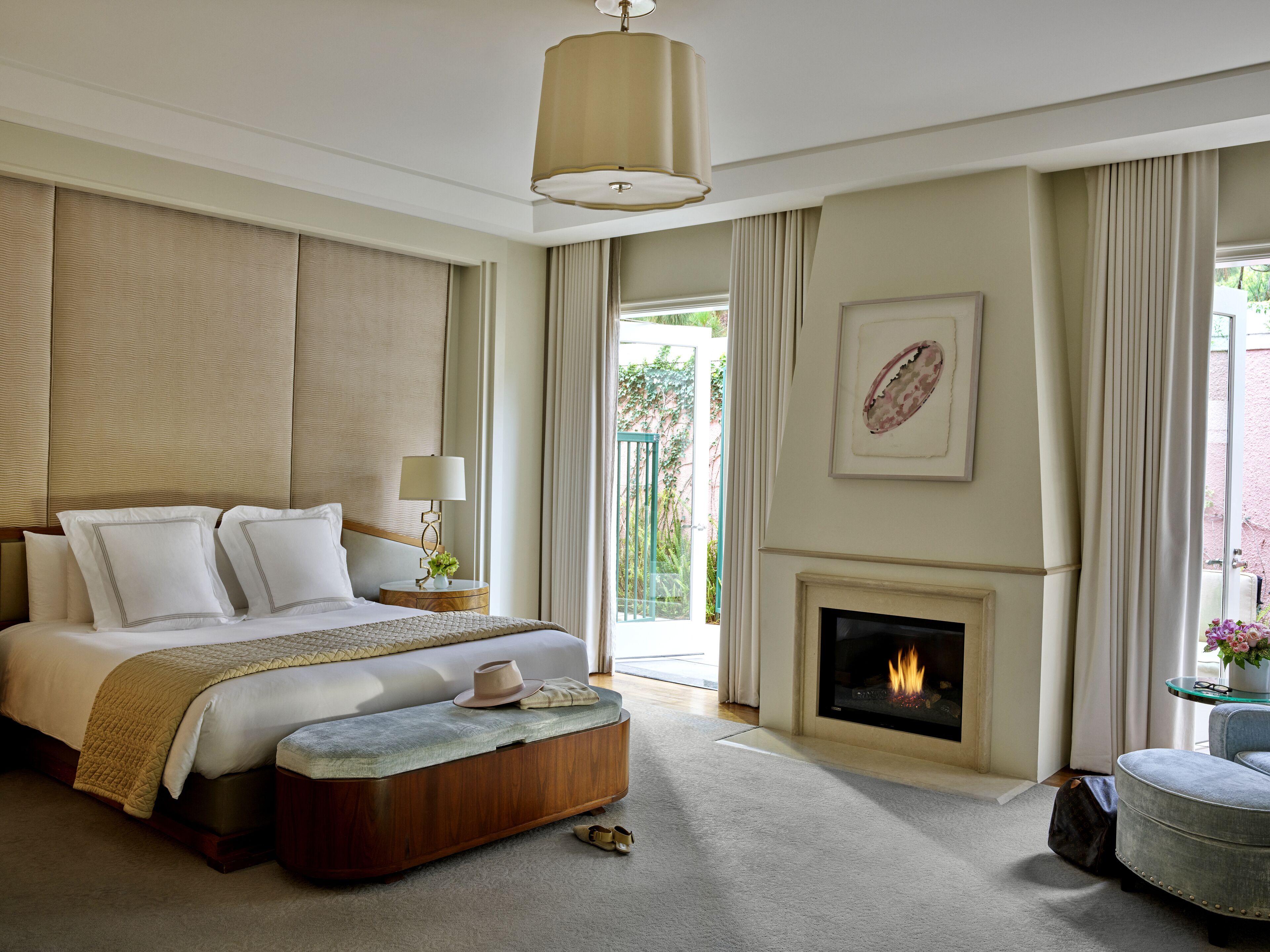 A spacious bedroom with a large bed, a bedside table with a lamp, a fireplace, and a view of greenery outside.