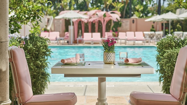 A poolside table with two chairs, a vase with flowers, and a view of a pool with lounge chairs and palm trees in the background.