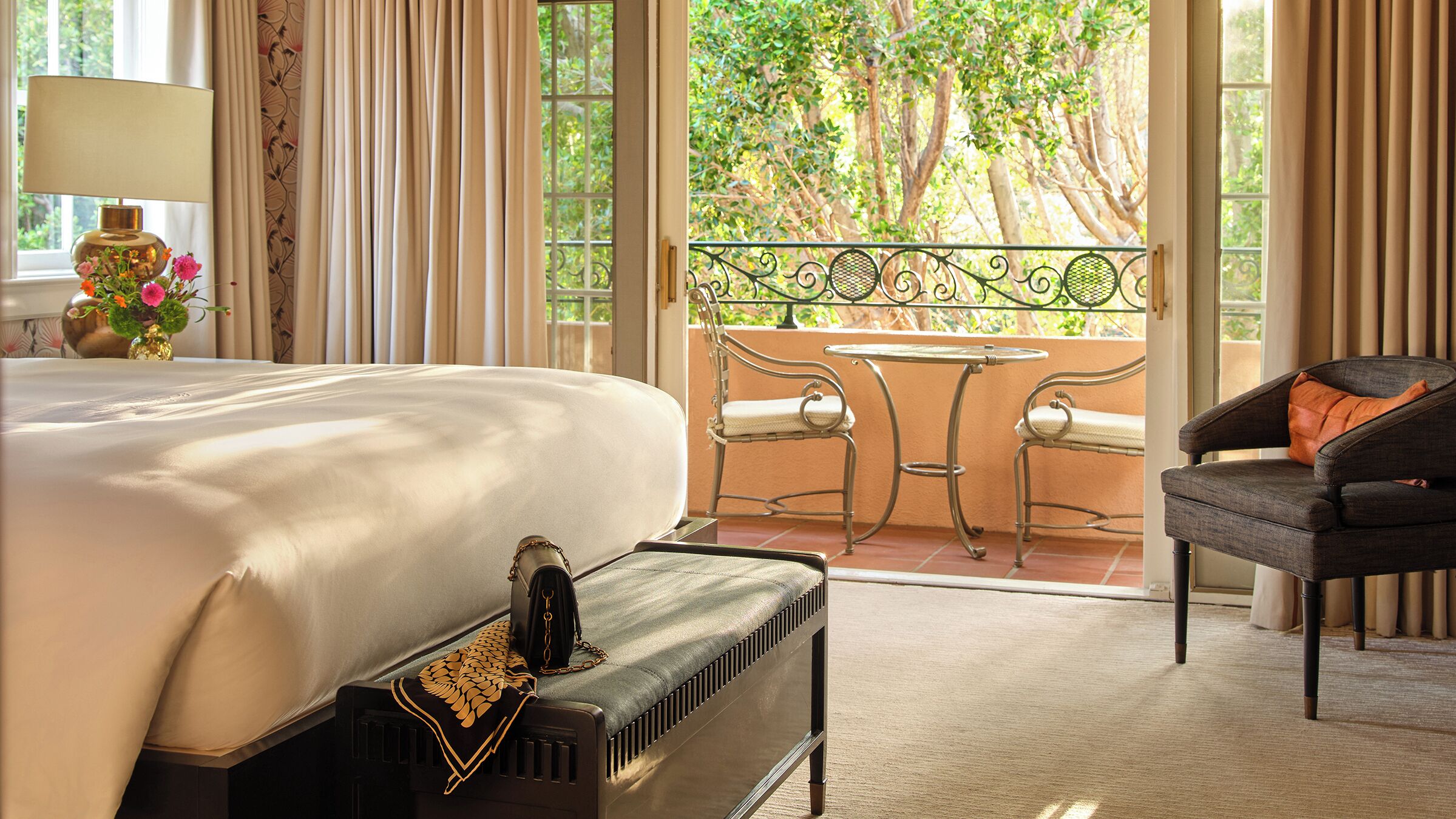A hotel room with a large bed, a bench with a decorative cushion, a table with two chairs, and a view of trees through the window.
