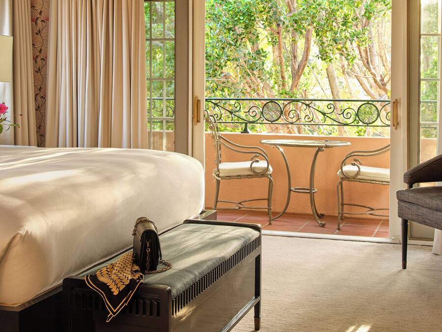 A hotel room with a large bed, a bench with a decorative cushion, a table with two chairs, and a view of trees through the window.