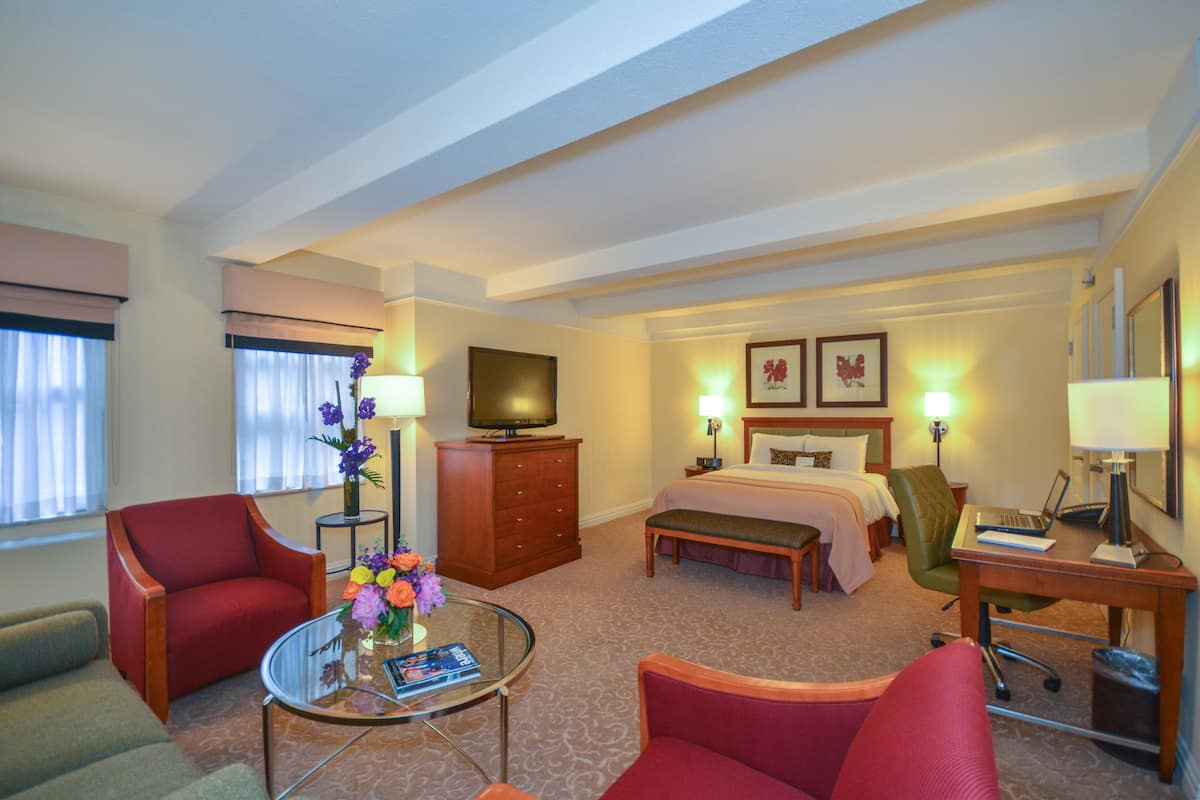 A hotel room with a bed, two chairs, a desk, a television, and a small table with flowers.