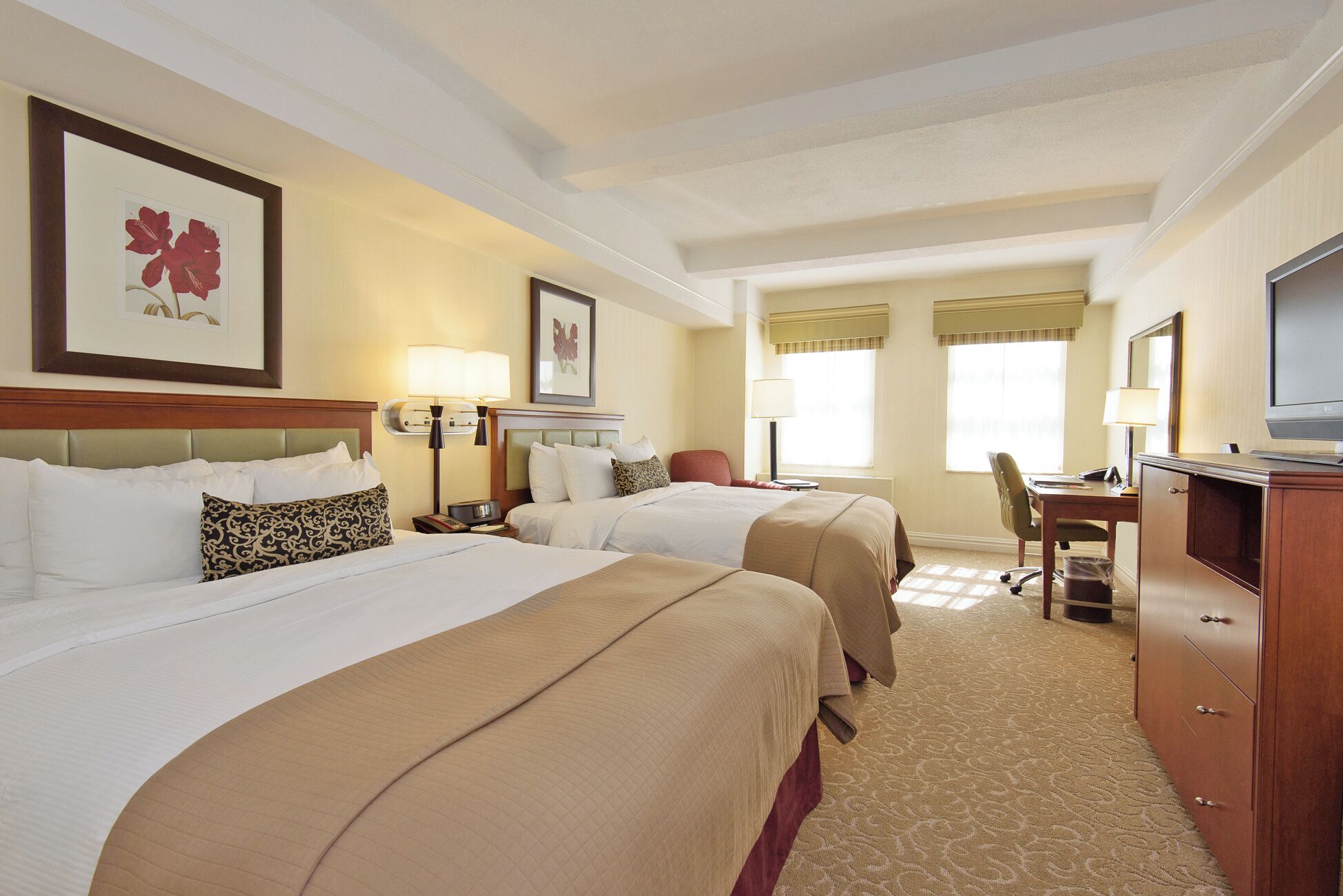 A hotel room with two beds, a desk with a chair, a TV, and framed artwork on the walls.