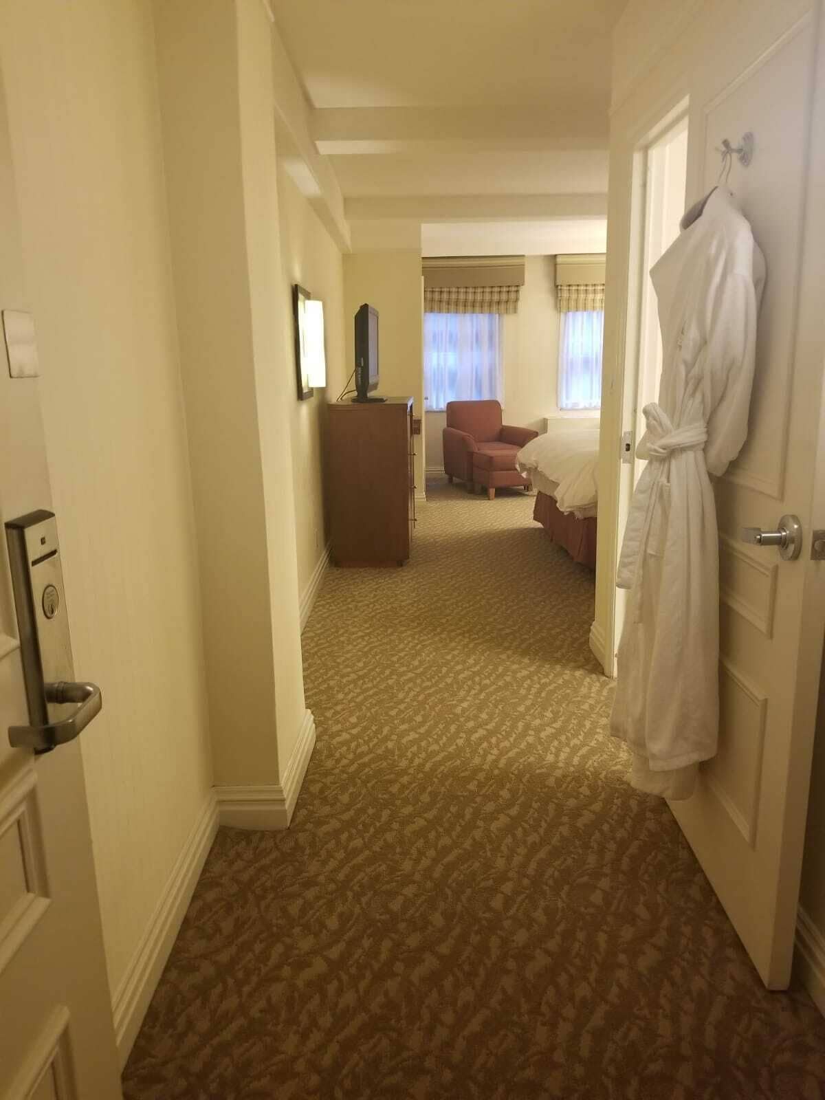 A hotel room with a carpeted floor, a bed, a chair, a desk with a television, and a robe hanging on the door.