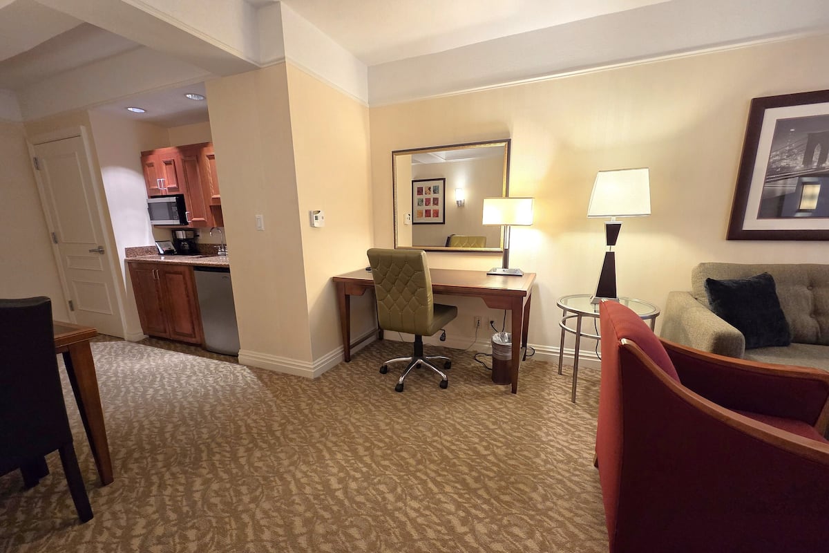 A hotel room with a kitchenette, a desk with a chair, a small round table, a sofa, and a lamp.