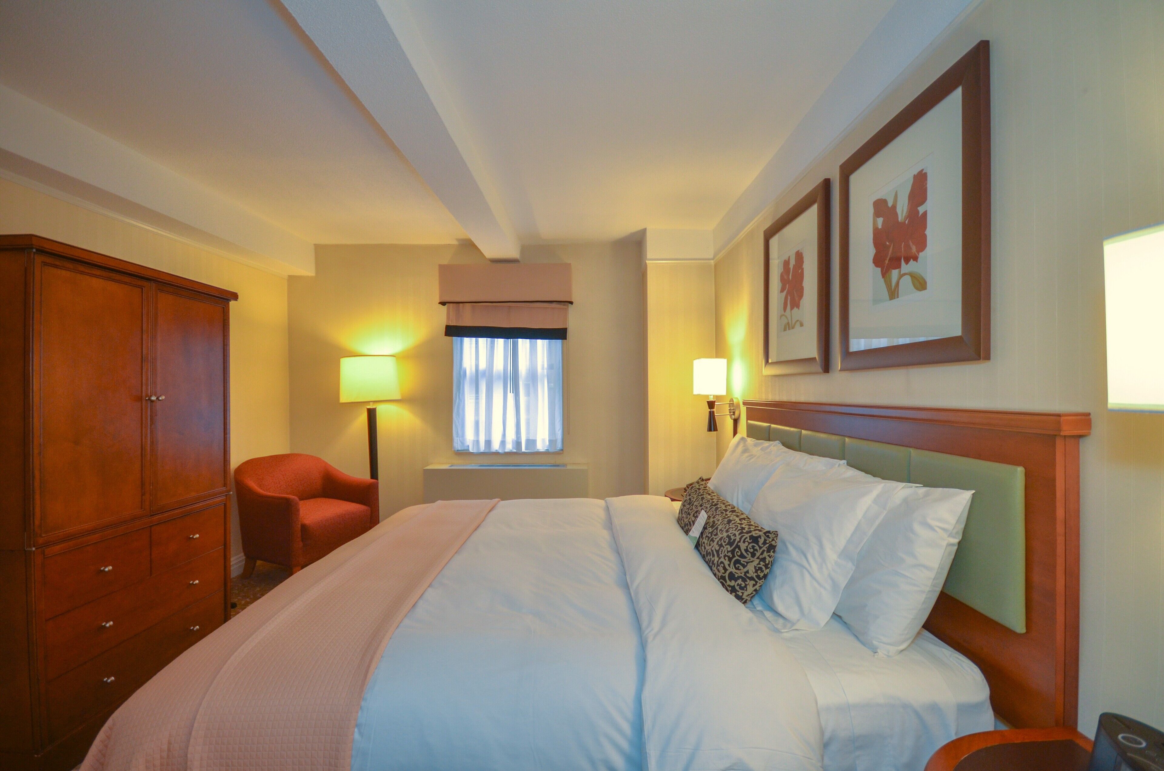 A hotel room with a bed, a wardrobe, a chair, two wall-mounted lamps, and framed artwork.