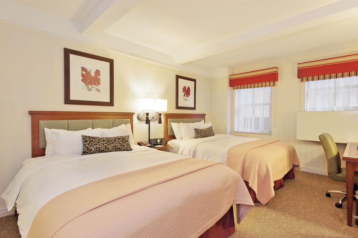 A hotel room with two beds, a desk, and a chair. There are framed pictures on the wall and a lamp on the desk.