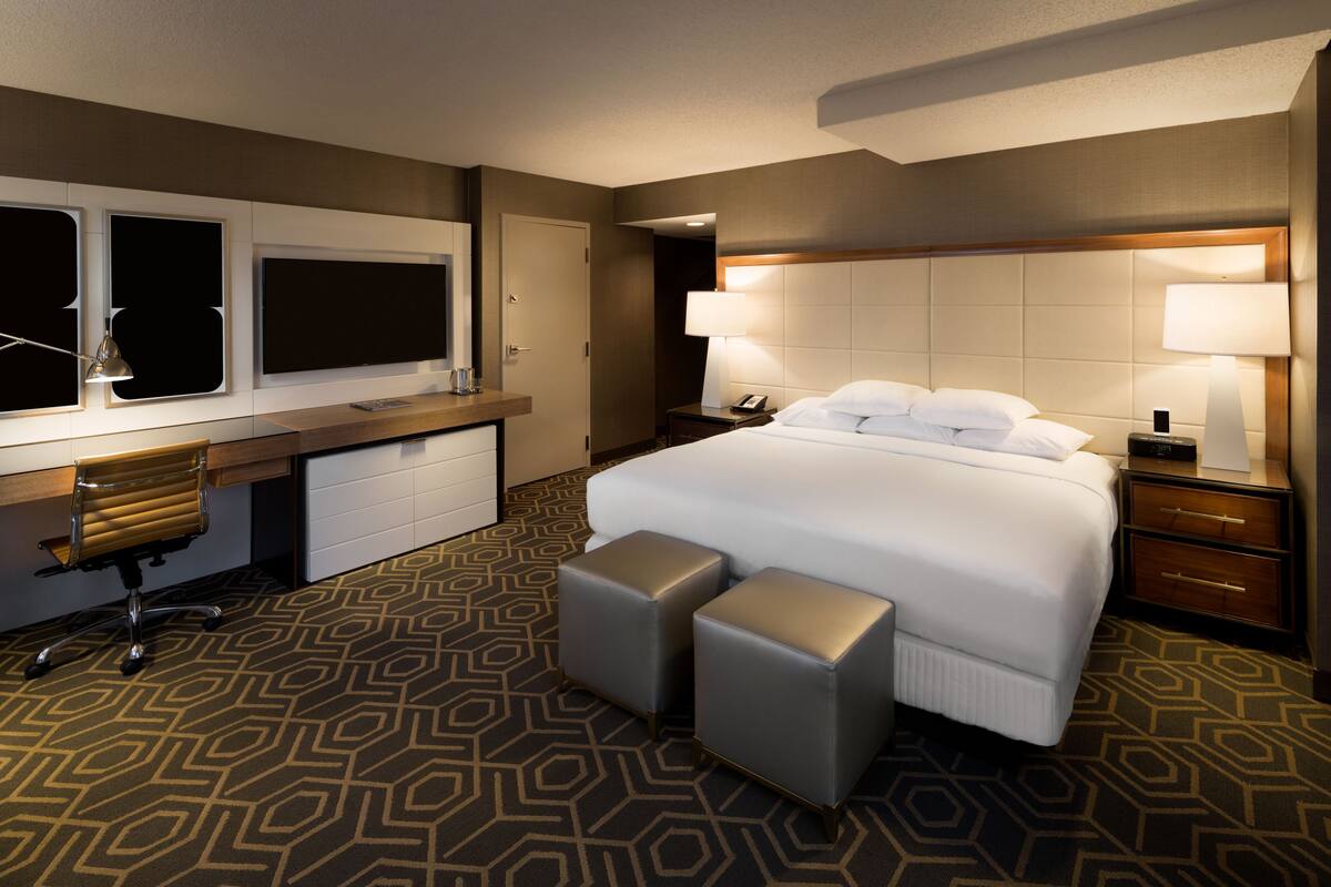 A hotel room with a large bed, a desk with a chair, a desk lamp, and a nightstand with a lamp.