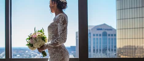 Une mariée, vêtue d’une robe de mariée en dentelle, tient un bouquet et se tient dans une pièce dotée de grandes fenêtres donnant sur un paysage urbain.