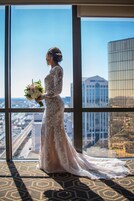 Una novia con vestido de encaje y un ramo de flores, de pie en una habitación con ventanas amplias que dan a una vista panorámica de la ciudad.