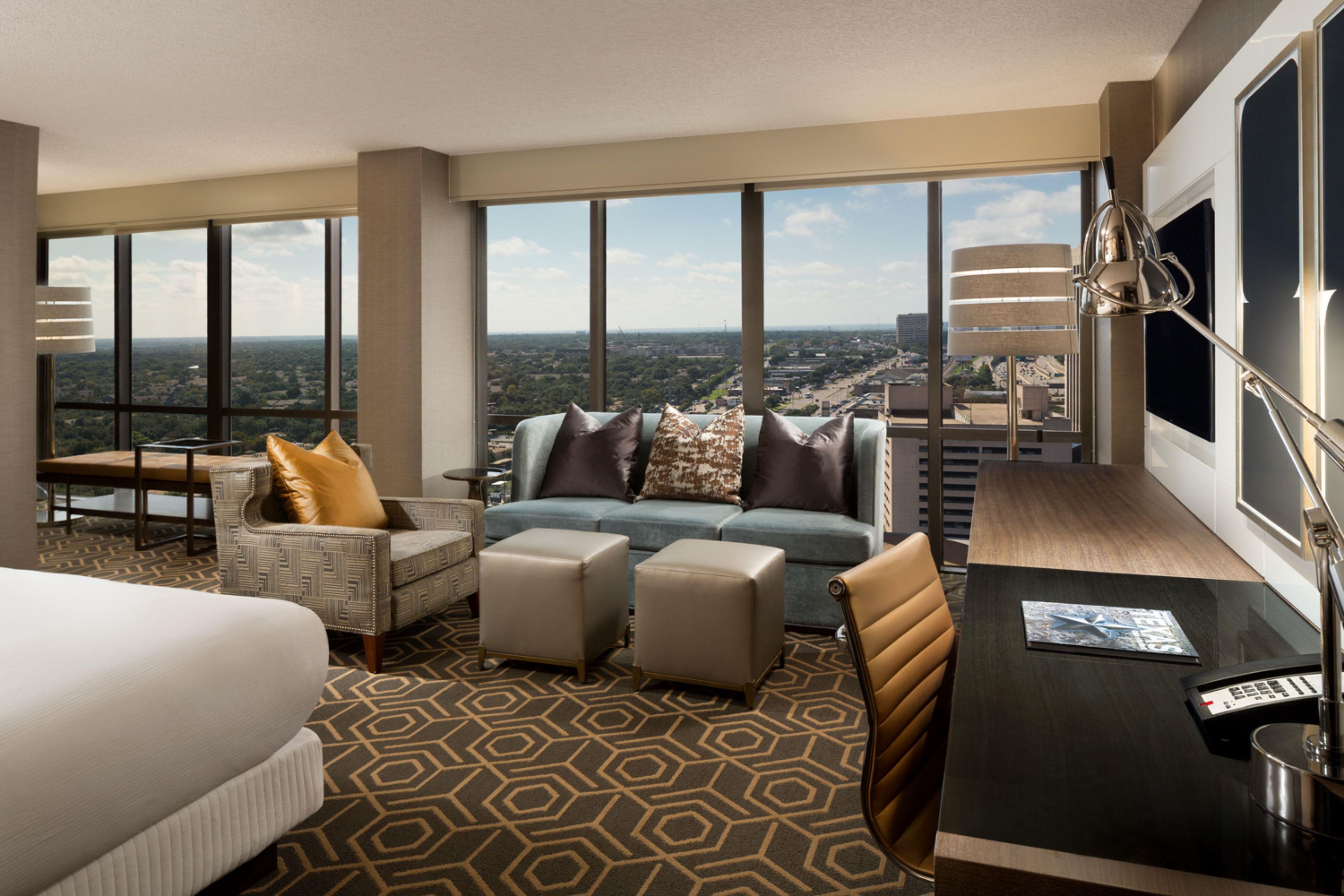 Ein Hotelzimmer mit einem großen Bett, einer Couch, zwei Hocker, einem Schreibtisch und einem Sessel. Große Fenster bieten Blick auf die Stadt.