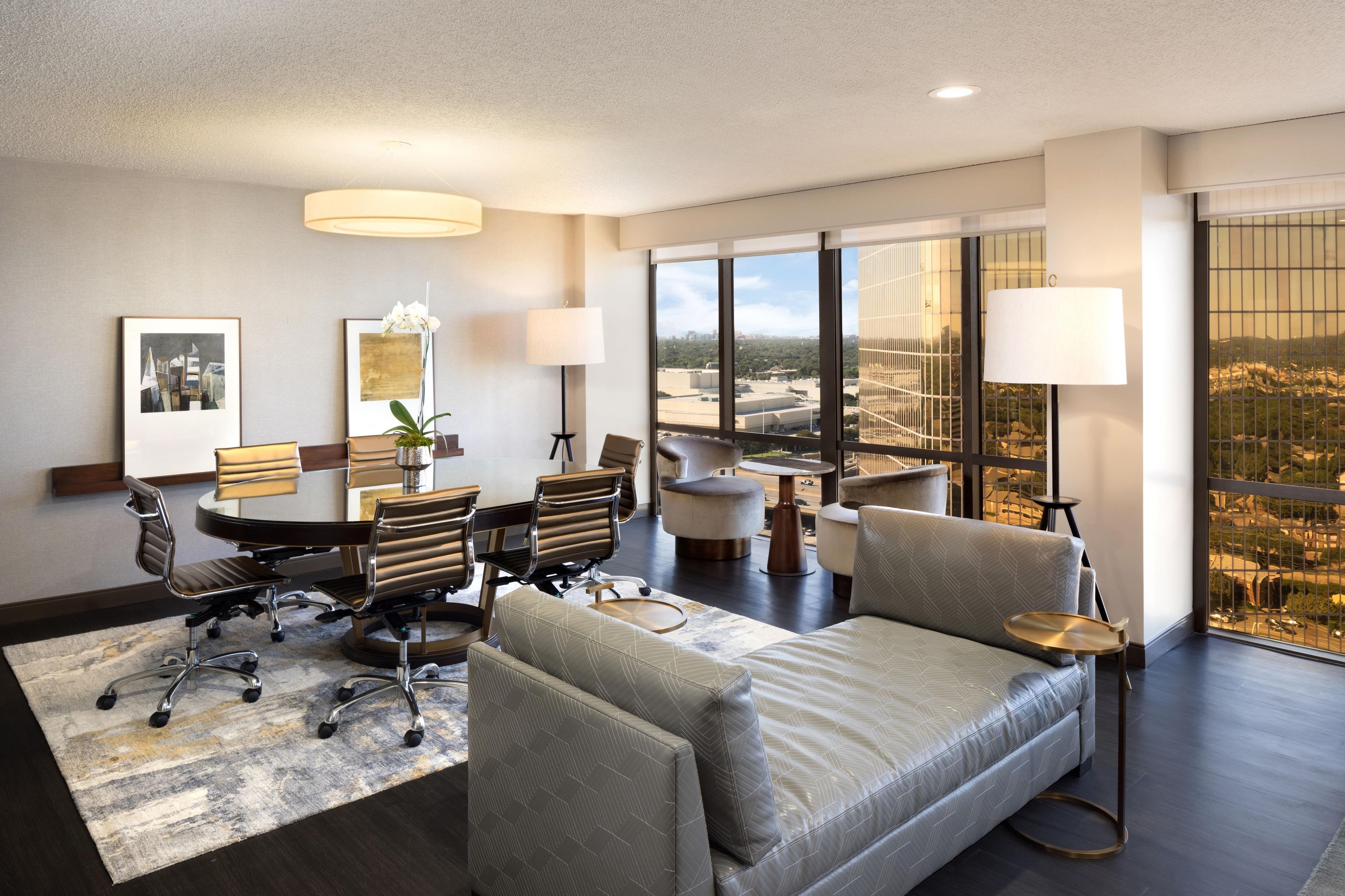 A modern hotel room with a large bed, a desk, two chairs, a sofa, and a view of the cityscape.