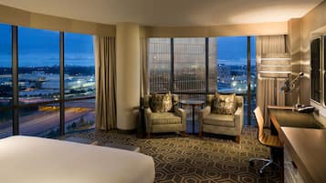 Habitación de hotel con una cama grande, dos sillones, una mesita, un escritorio con silla y un ventanal con vistas a la ciudad.
