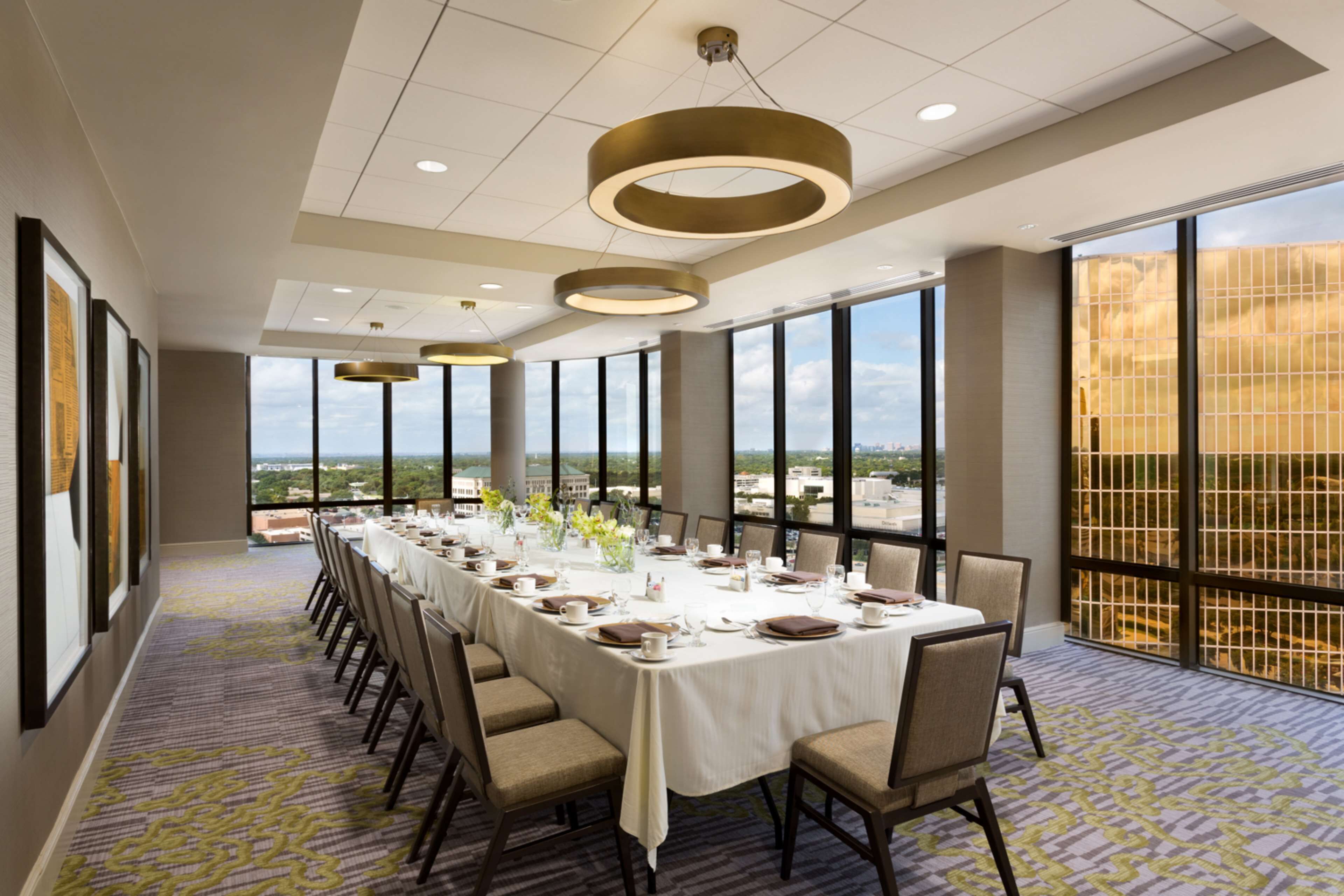 A conference room with a long table set for a meeting, large windows offering a city view, and modern pendant lighting.