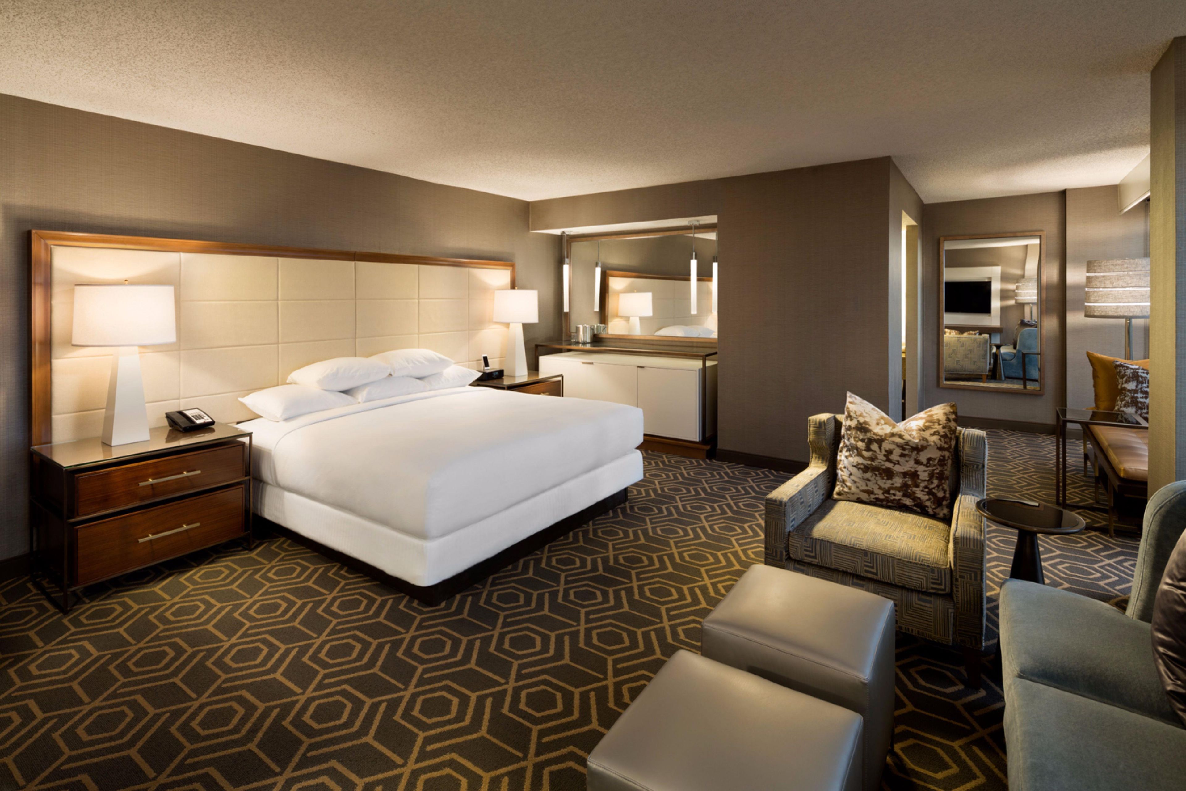 A hotel room with a large bed, bedside tables, a desk, a chair, a sofa, and a mirror.