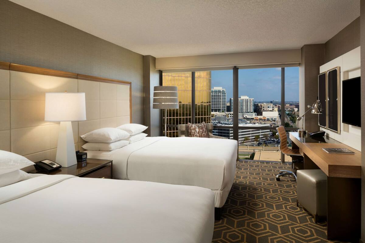 A hotel room with two beds, a desk, a TV, and a large window with a city view.