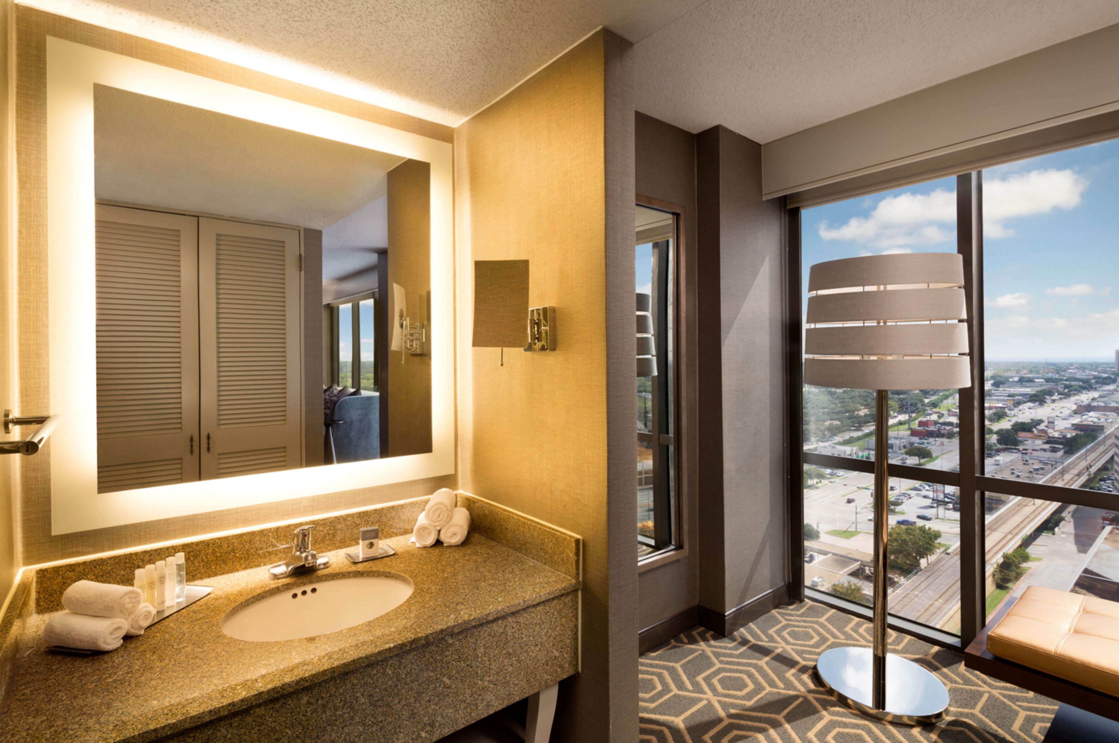 Un baño de hotel con un espejo grande, encimera de granito y vistas de la ciudad a través de un ventanal.
