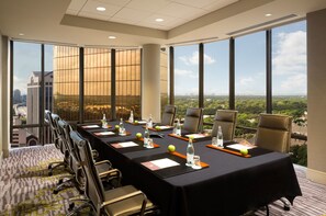 Un salón de conferencias con una larga mesa, sillas, botellas de agua y vistas a la ciudad a través de grandes ventanales.