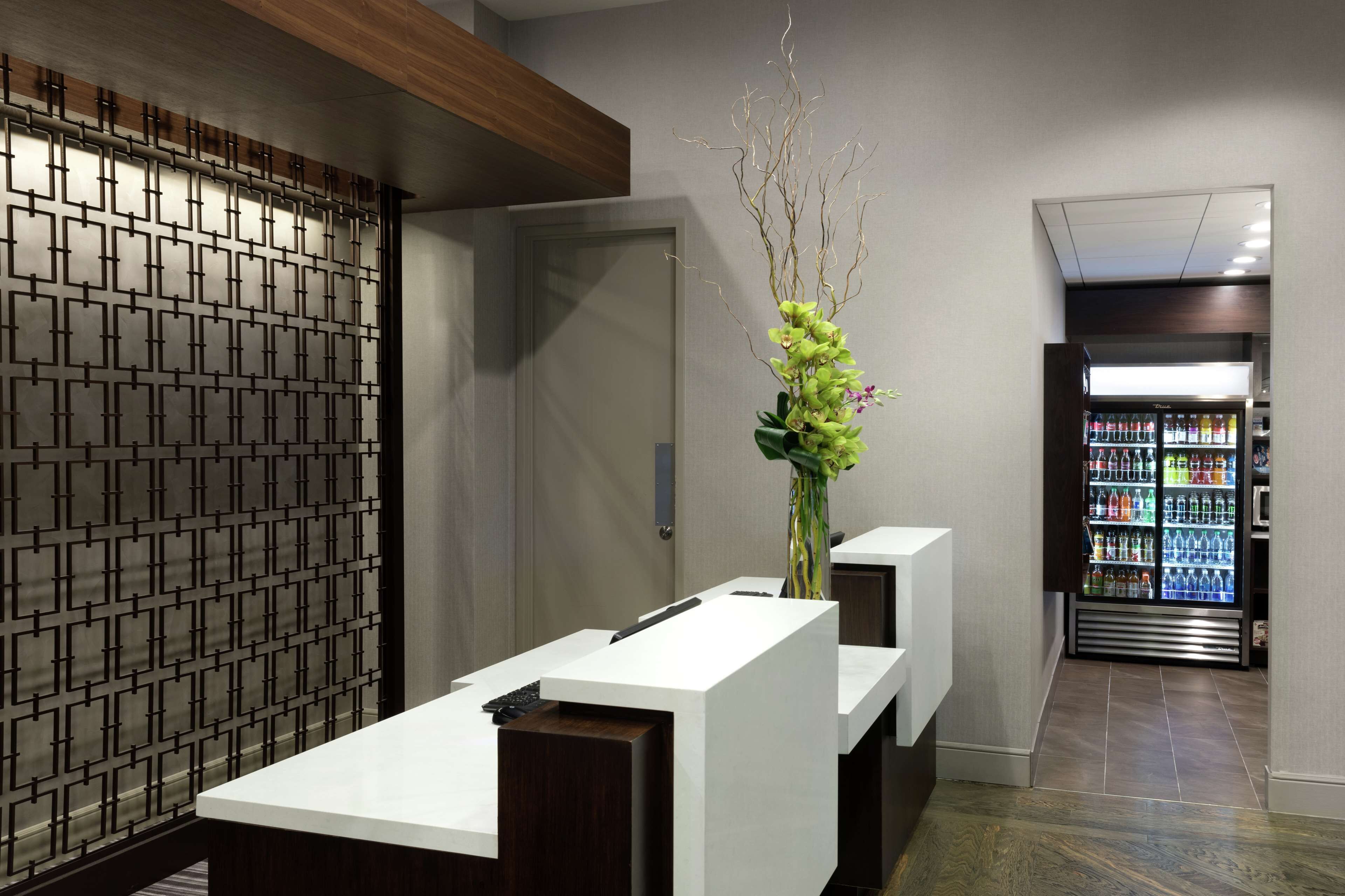 A reception area with a modern design, including a reception desk, a decorative vase with greenery, and a refrigerator stocked with beverages.
