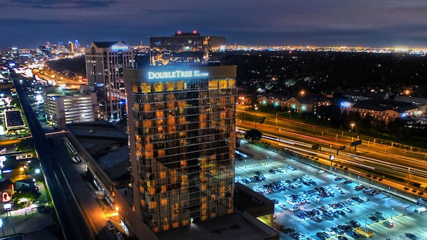 Een hoog hotel met een opvallend “DOUBLE TREE BY HILTON” bord, gelegen in een levendige stedelijke omgeving met zichtbaar verkeer en parkeerplaatsen.