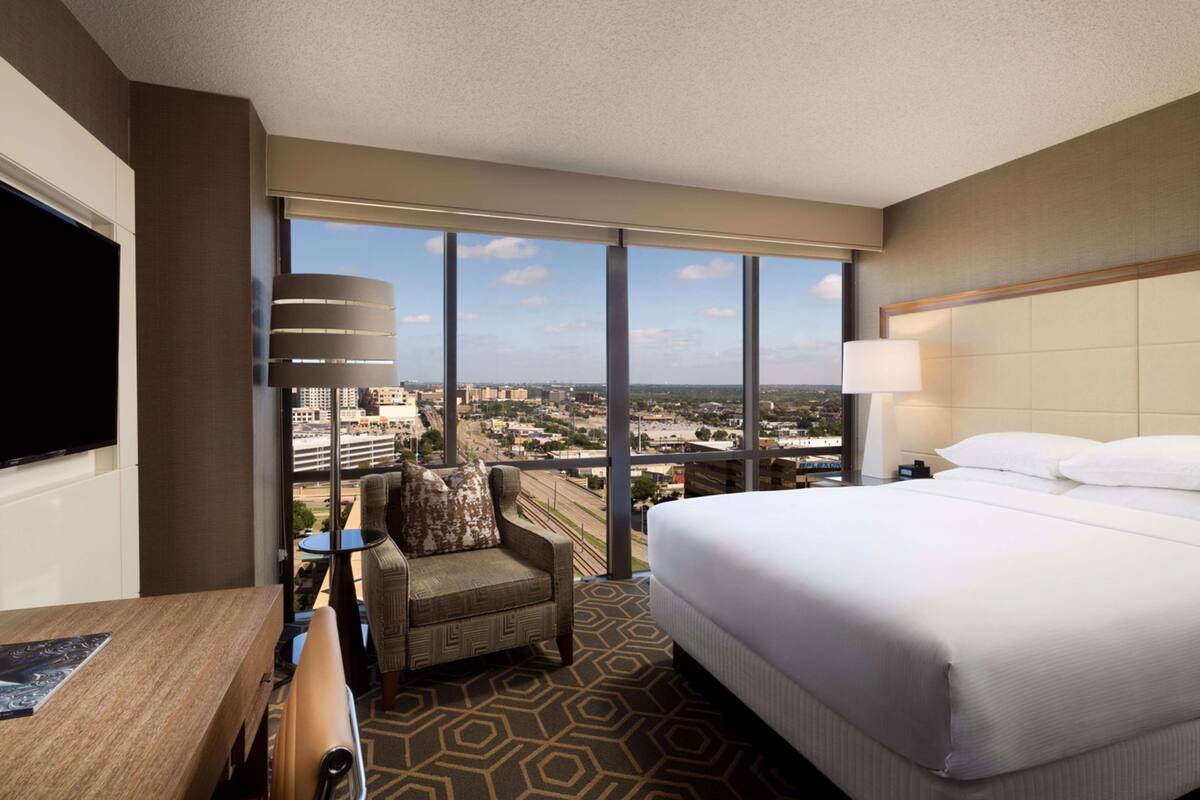 A hotel room with a large bed, a chair, a desk, and a TV. The room has large windows offering a city view.