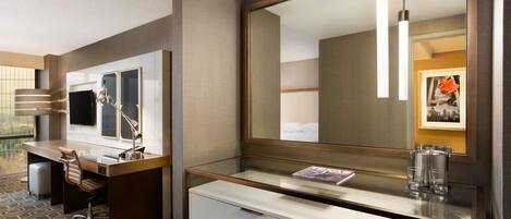 Habitación de hotel con escritorio, silla y un espejo grande. Hay un televisor en la pared y una ventana que ofrece vistas al exterior.