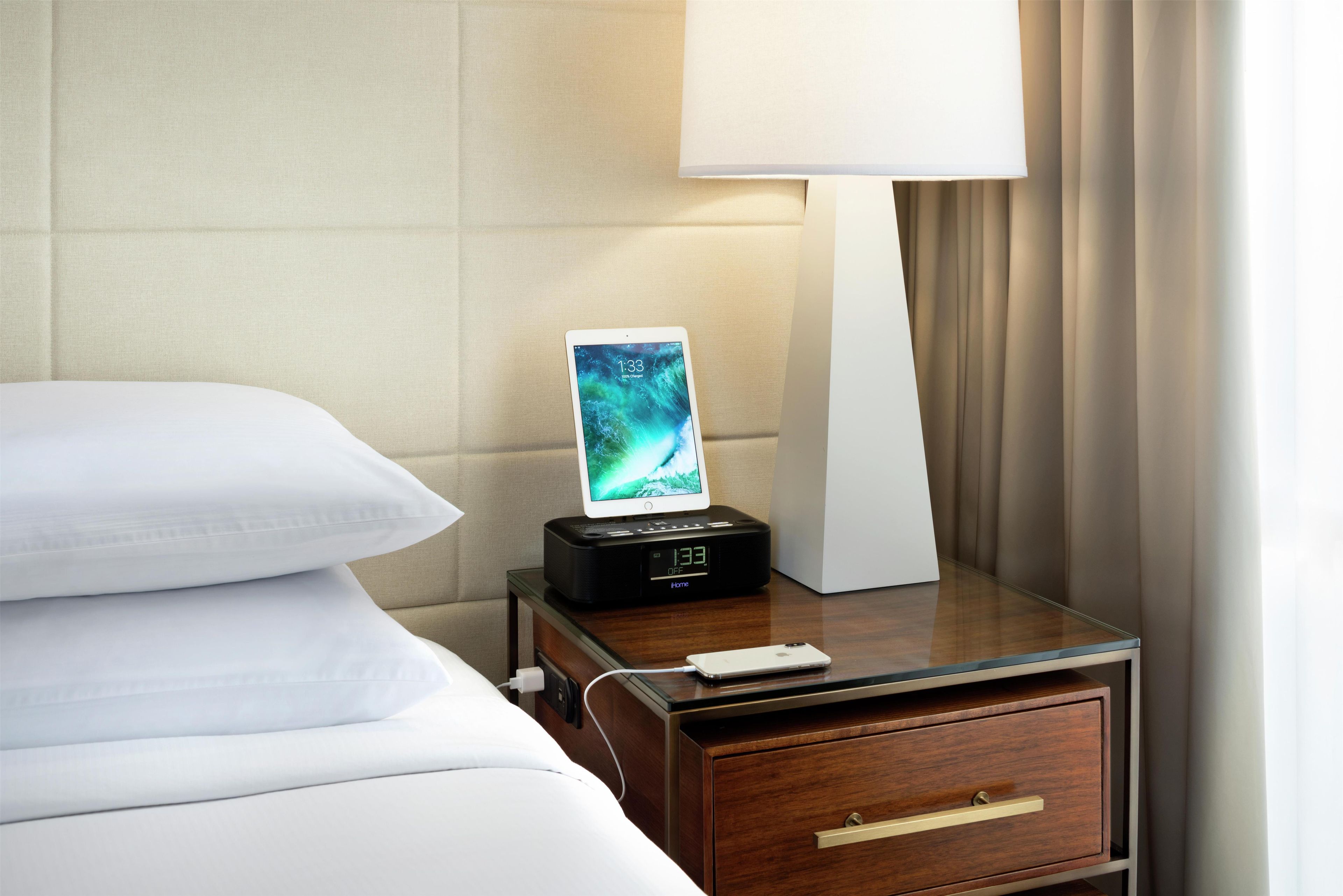 Habitaci ón de hotel impecable con cama, mesita de noche con lámpara, reloj digital y teléfono inteligente cargando.