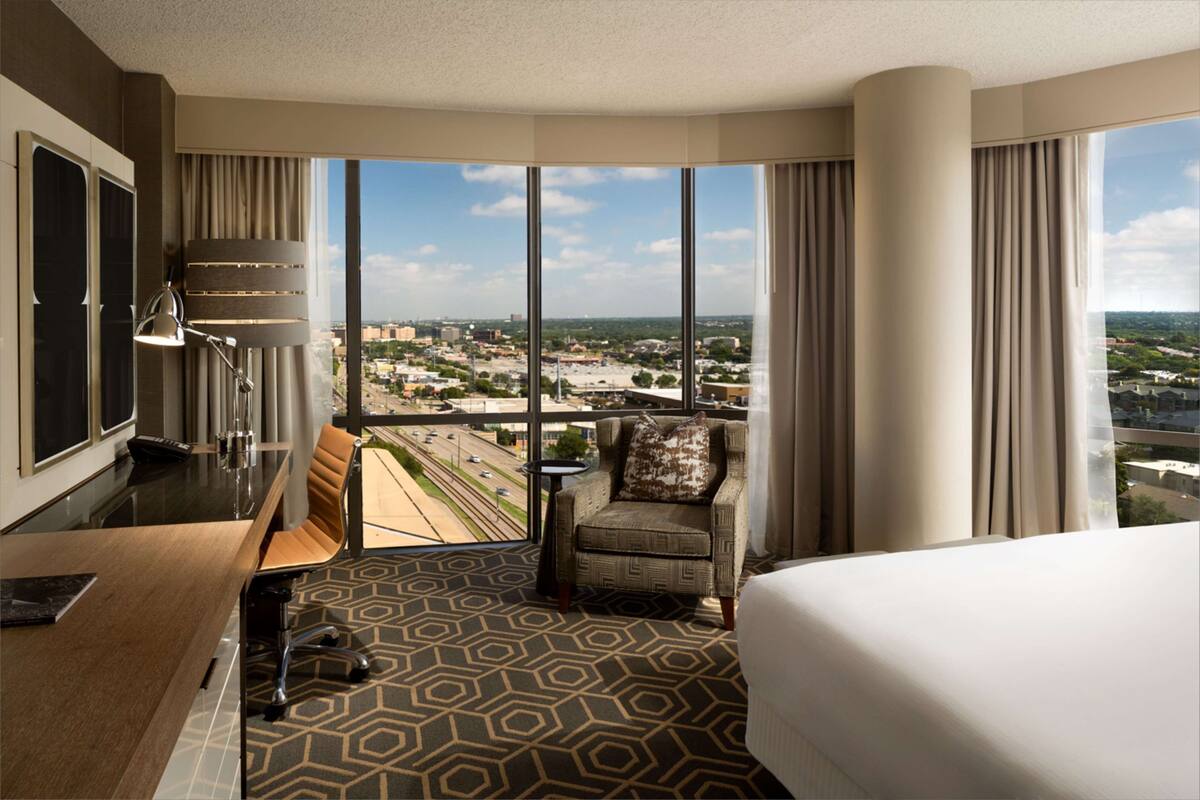 A hotel room with a large window, a desk, a chair, and a bed.