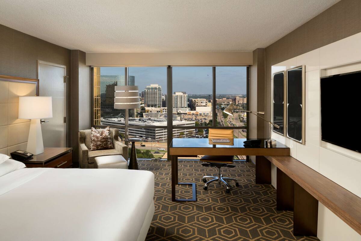 A hotel room with a large bed, a desk, and a view of the cityscape through large windows.